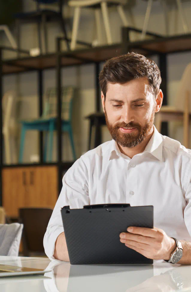 A well dressed man doing work on an tablet in furniture store, display of chairs in the background.