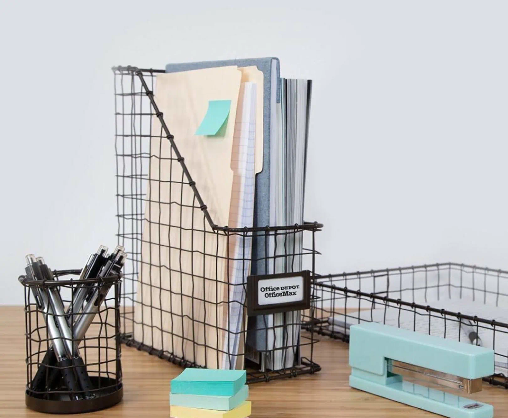 Organized office desk with pen holder, sticky notes, magazine holder with files, stapler, and paper tray. The label on the magazine holder says "office depot, office max"