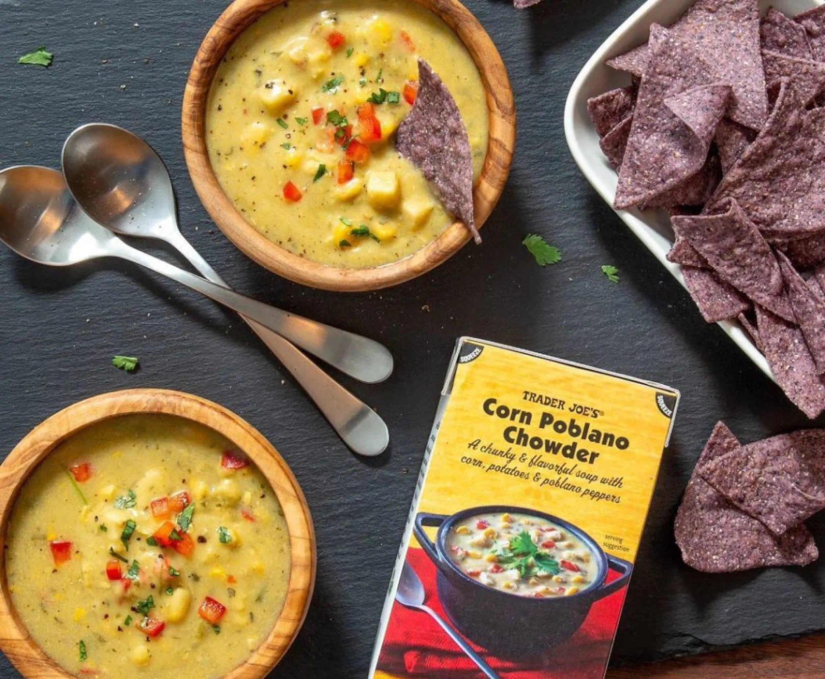Two bowls of corn chowder, a box of Trader Joe's Corn Poblano Chowder, two spoons, and a plate of blue corn tortilla chips.