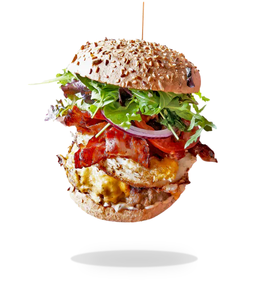 Fancy burger with a sesame bun in the air showing layers of lettuce, bacon, tomatoes and patty.