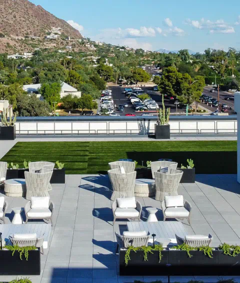 Rooftop terrace with modern lounge chairs, green grass, potted plants, overlooking a parking lot, residential area, and distant mountains.