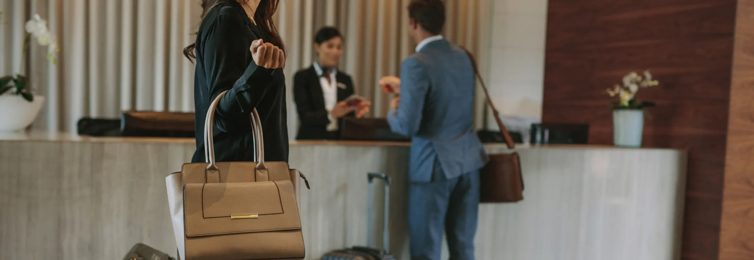 Woman with a beige purse walks towards reception at a nice hotel. A man is in front of her at the front desk with his luggage checking in.