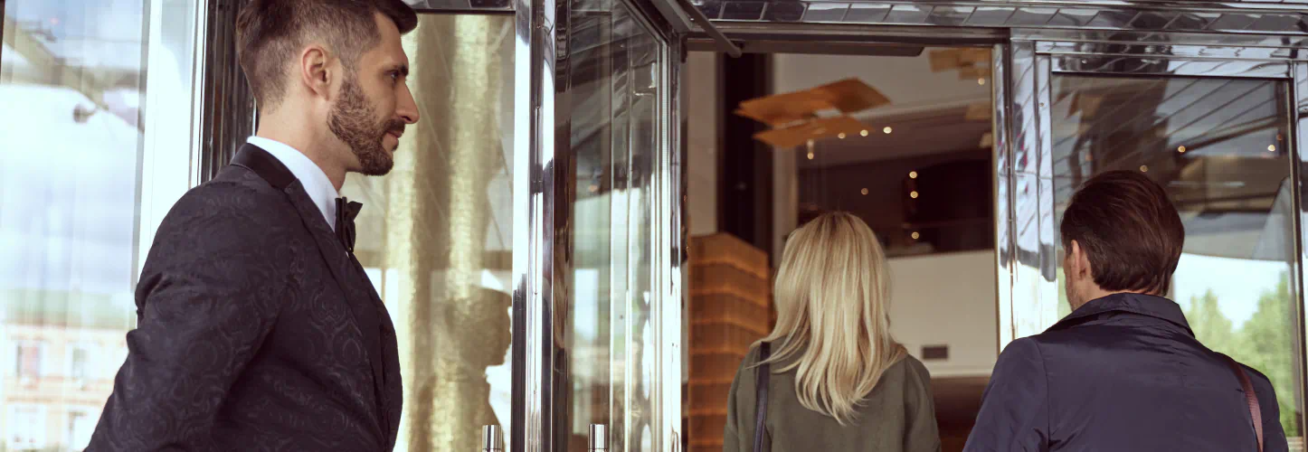 A doorman holds the entrance open for a couple wearing dark overcoats.