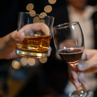 Two people toast. One is drinking whiskey on the rocks, the other is drinking wine.