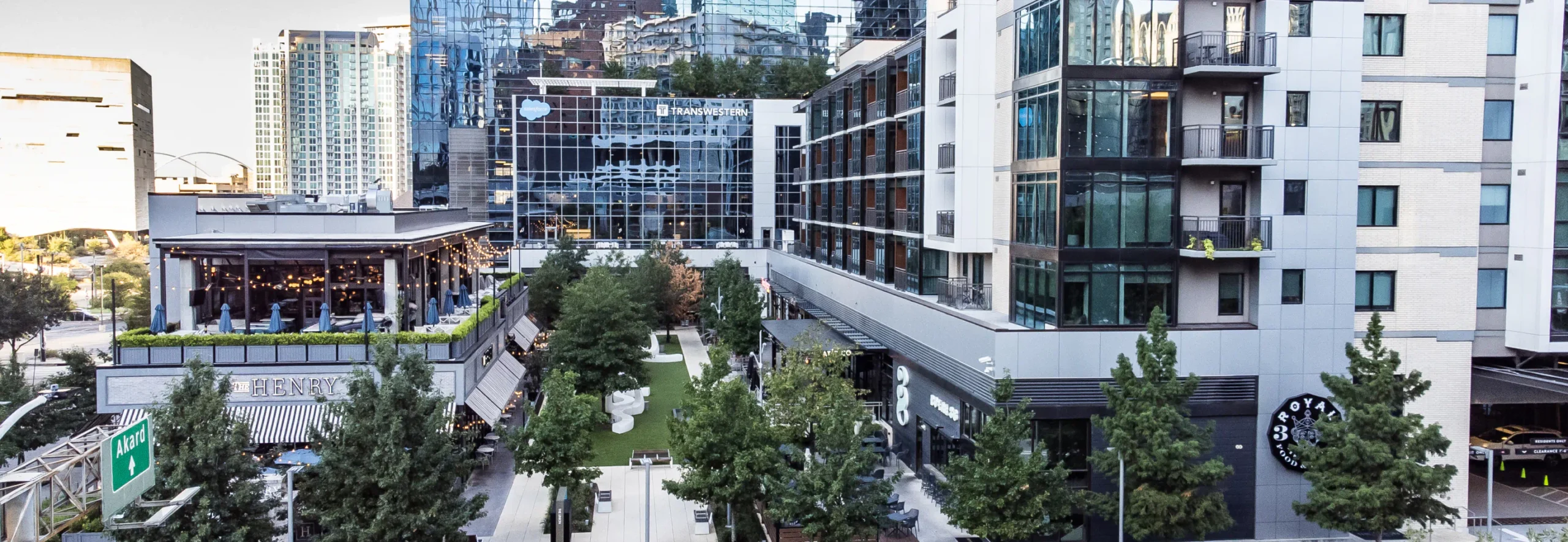 Aerial view of an outdoor space surrounded by luxury restaurants and apartments. Landscaping and trees fill the outdoor space and there is a rooftop bar on one of the buildings.