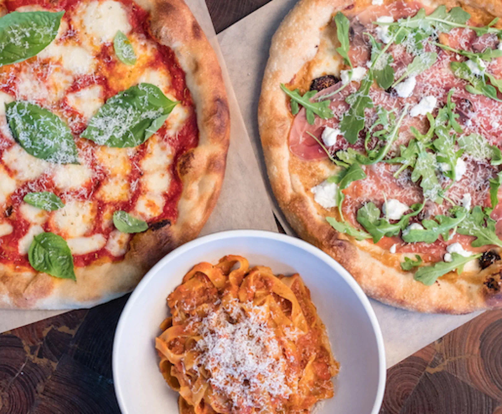Pizzas and pasta sit on a wooden table. They include a margarita pizza, a fig and prosciutto pizza, and a plate of pappardelle pasta.