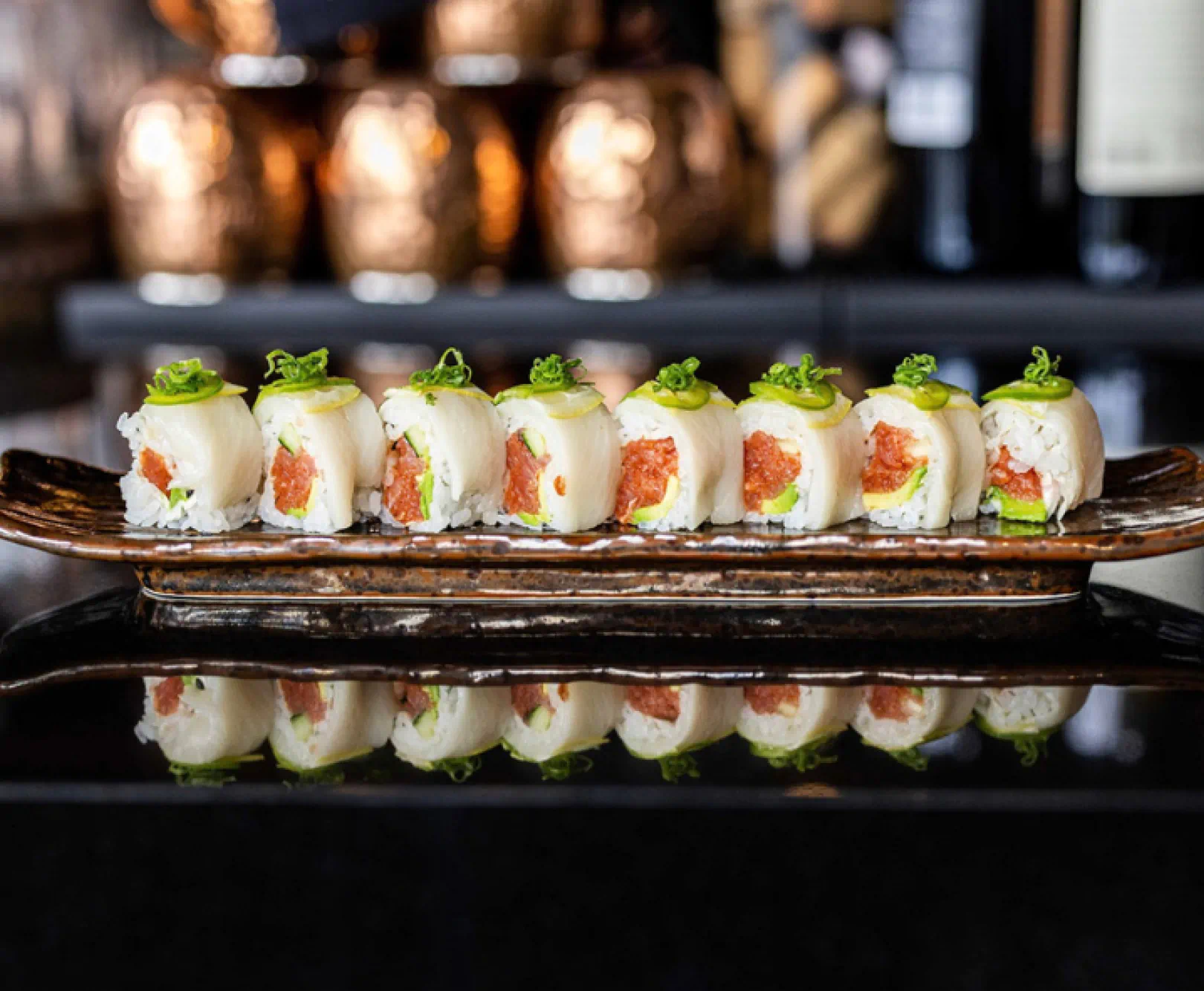 A sushi roll sits on a long plate, topped with sliced jalapeños and green onions.