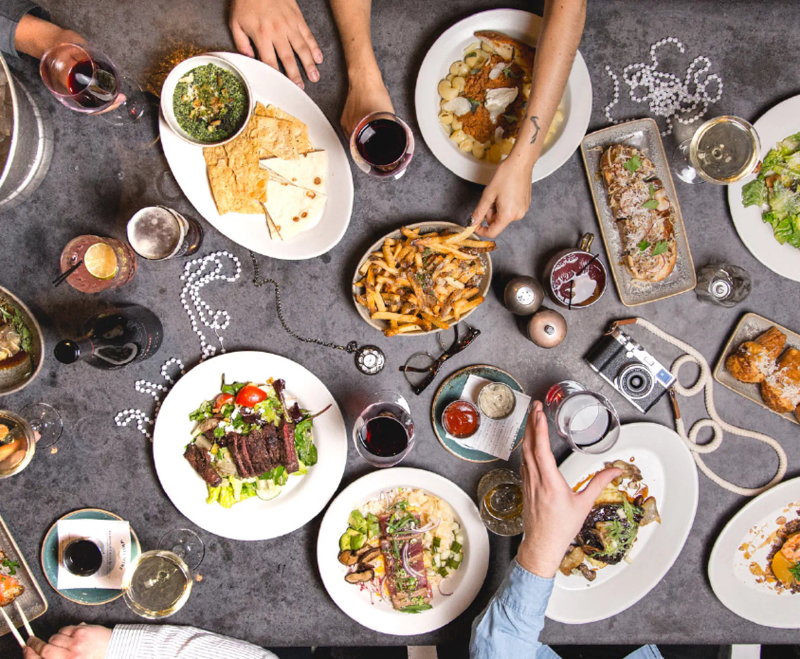Hands grab at a huge spread of food sitting on a metal table. It includes fries, quesadillas, steak salad, and bruschetta.