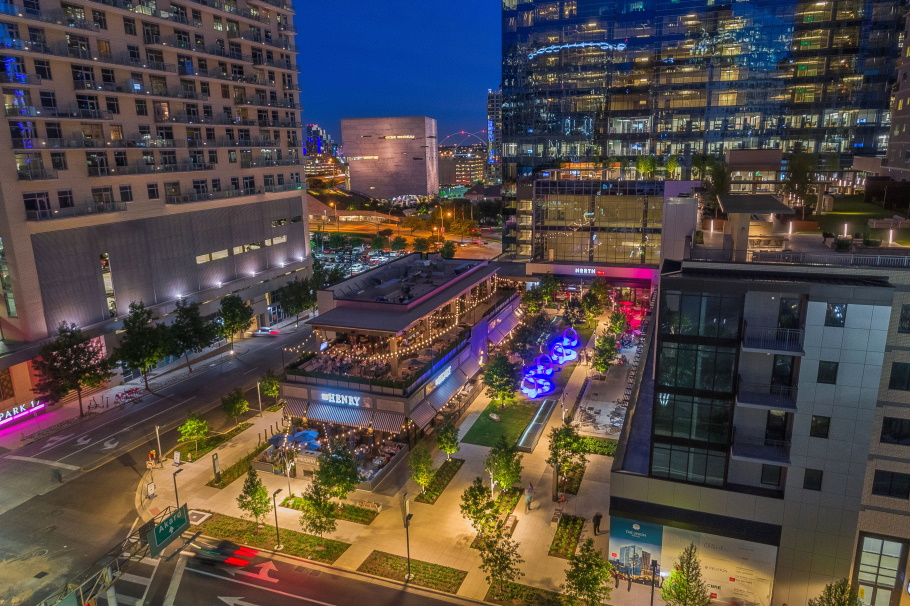 Aerial view of an outdoor space at night surrounded by luxury restaurants and apartments. Landscaping and trees fill the outdoor space and there is a rooftop bar on one of the buildings.