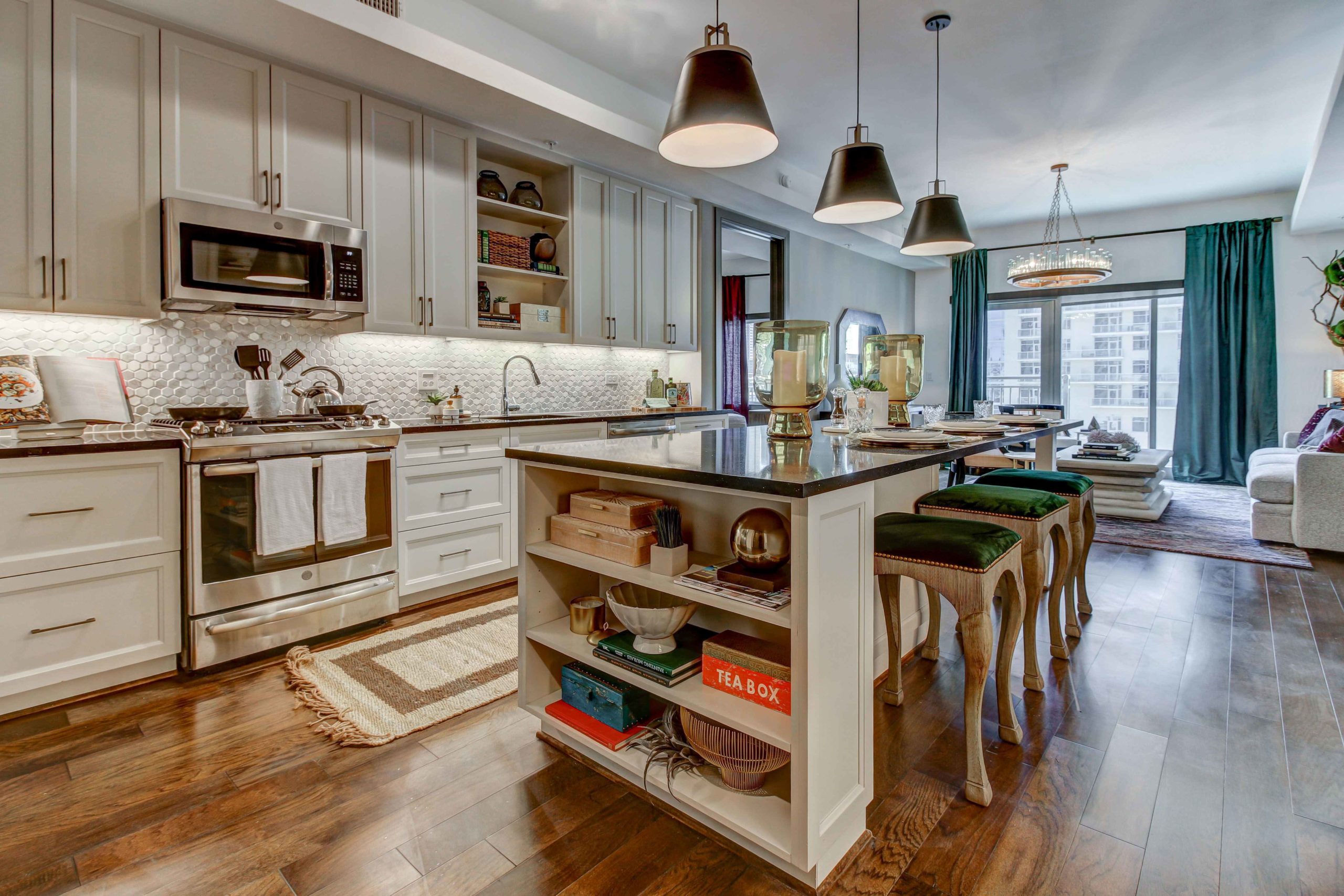 Luxury kitchen with a large island, white cabinets, new appliances, open shelving, and large pendant lights. The floors are wood and in the background there is a sitting area with a large window.