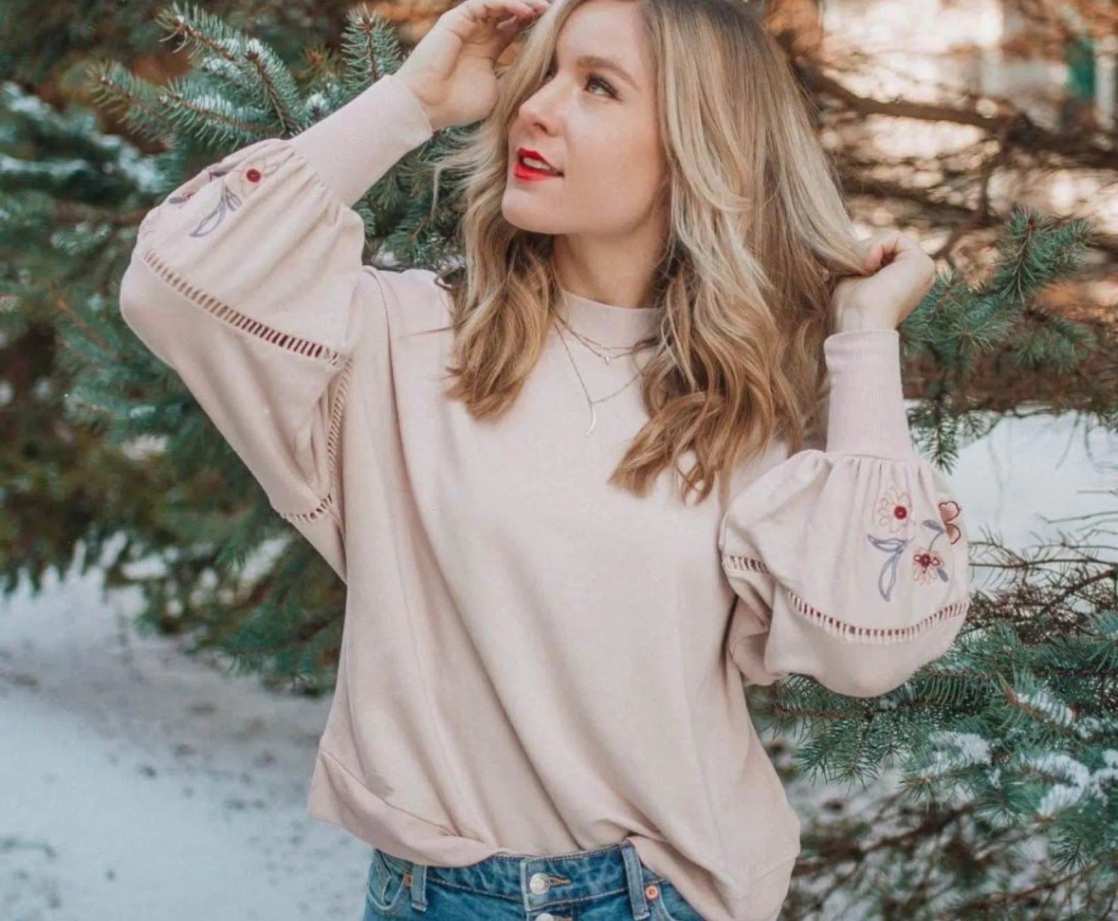 A girl with blond hair walks through snowy evergreens wearing a pink, flowy sweater and jeans.