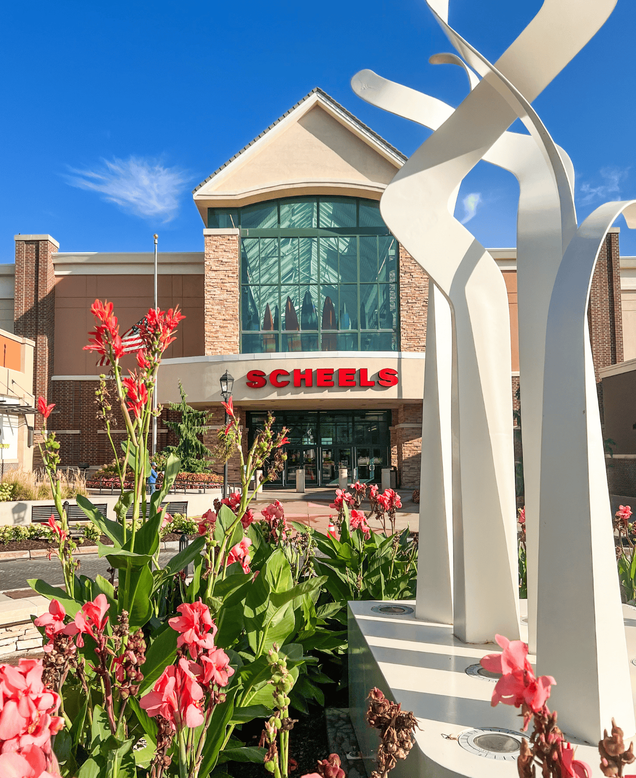 "Scheels" store front at Village Pointe. A white sculpture sits in the center of a roundabout adorned with plants with pink flowers in the foreground.