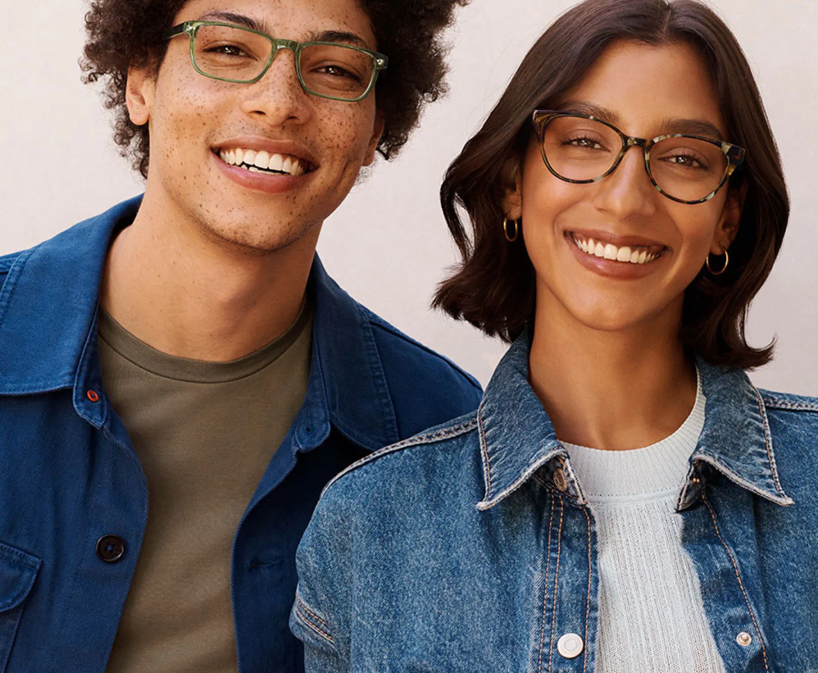 A man and woman wearing denim coats smile while wearing green and tortoise shell glasses.