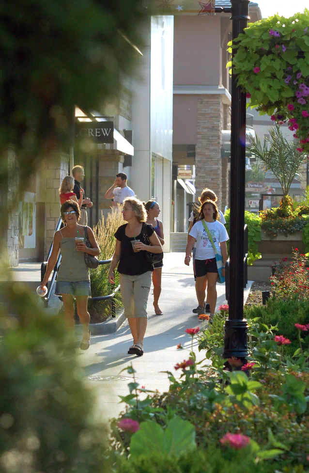 A group of shoppers walk in front of the store fronts of Village Pointe. Green plants with pink flowers line the walkway.