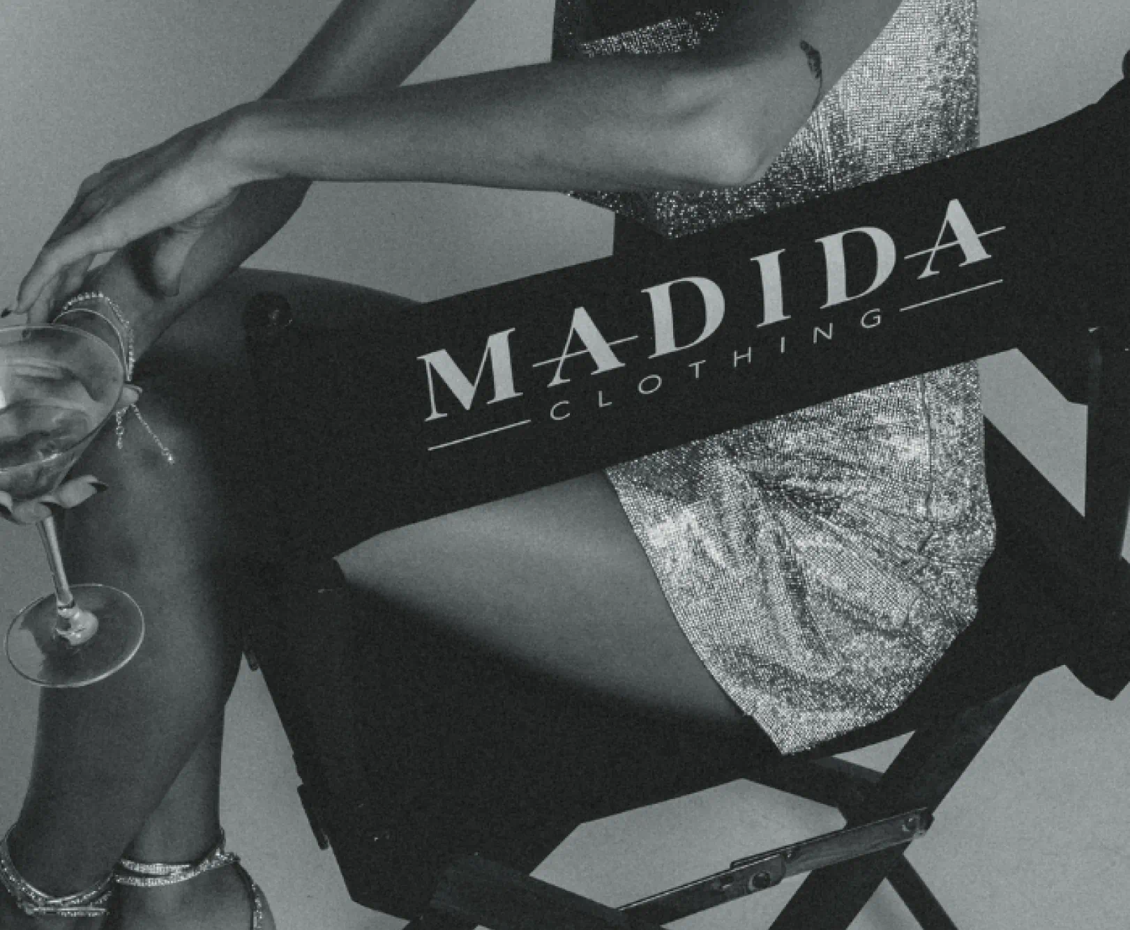 A women in a sparkly dress holds a martini while sitting in a director's chair with the Madida clothing logo on the back.