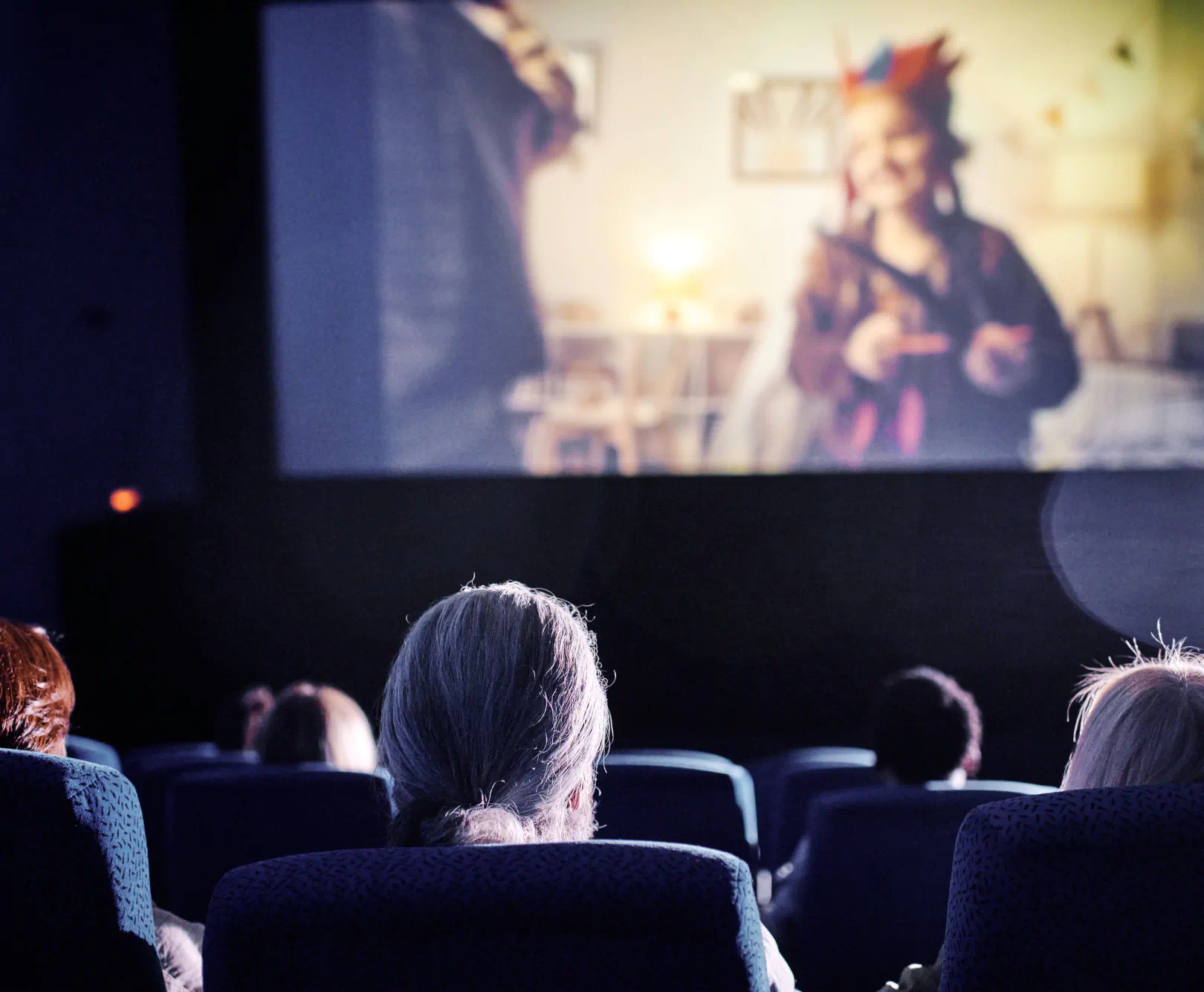 An audience watches a film in a dark theater. The actor on the screen wears a multi-colored hat.