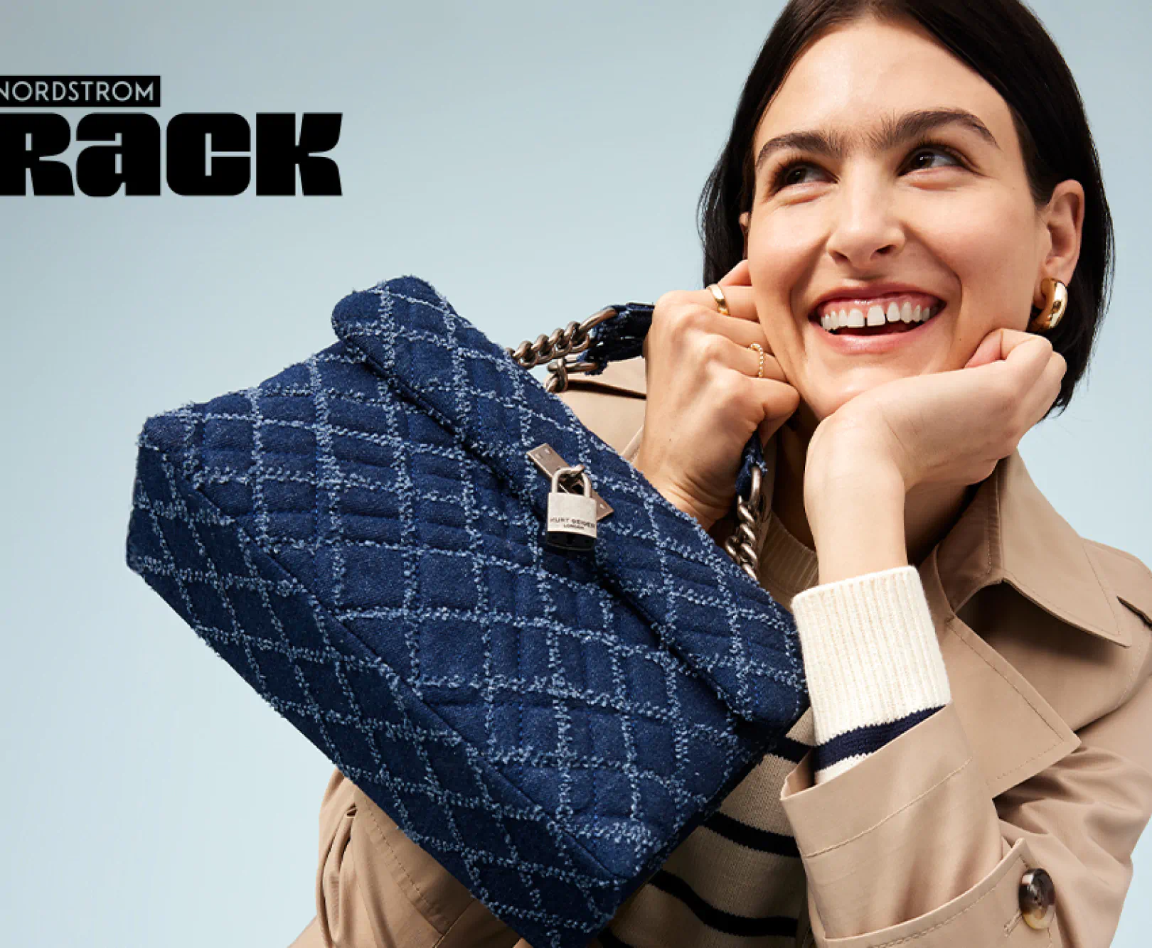 A woman in a tan overcoat smiles as she shows off her navy blue purse.