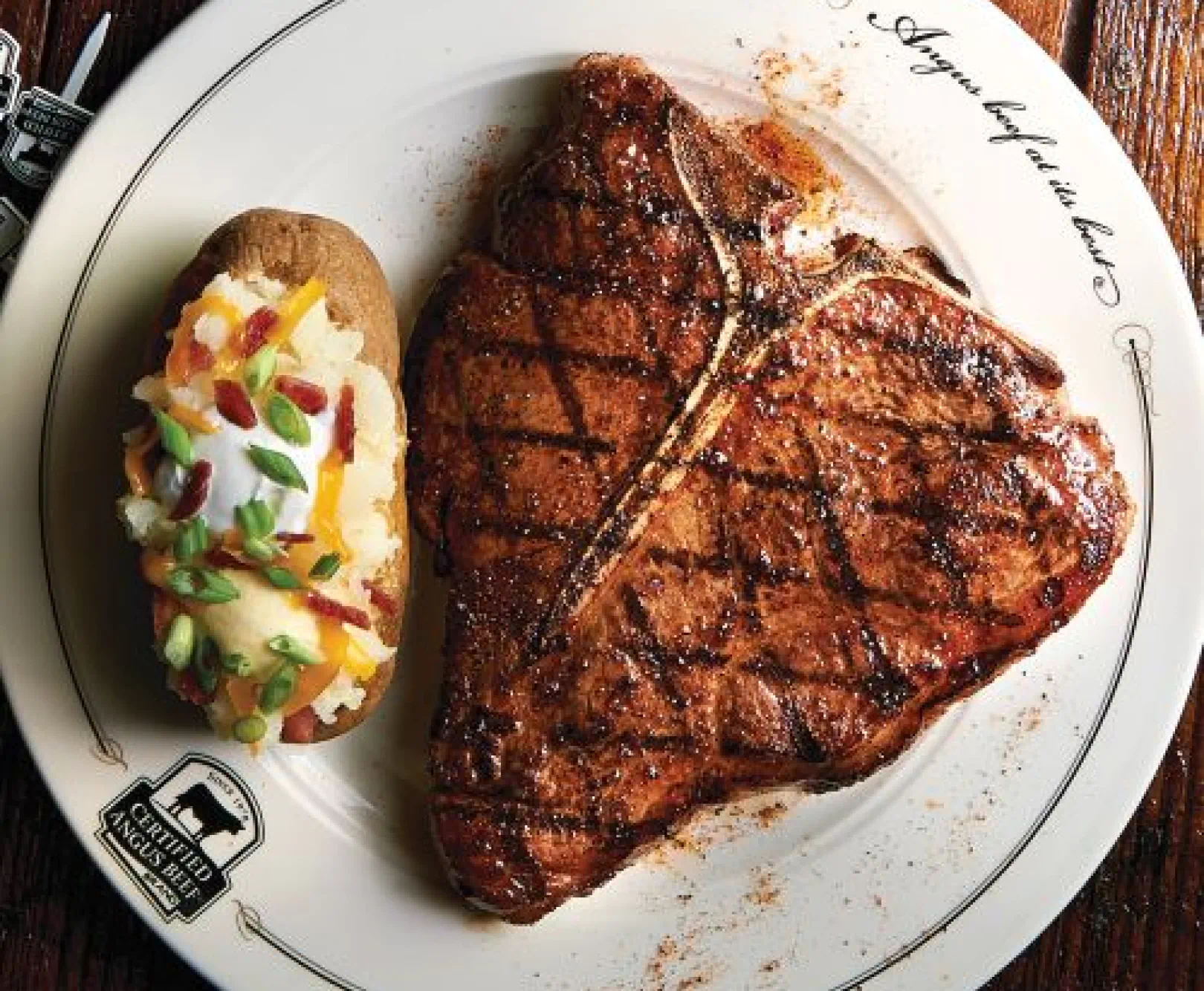 A T-bone steak and loaded baked potato sit on a white dinner plate.