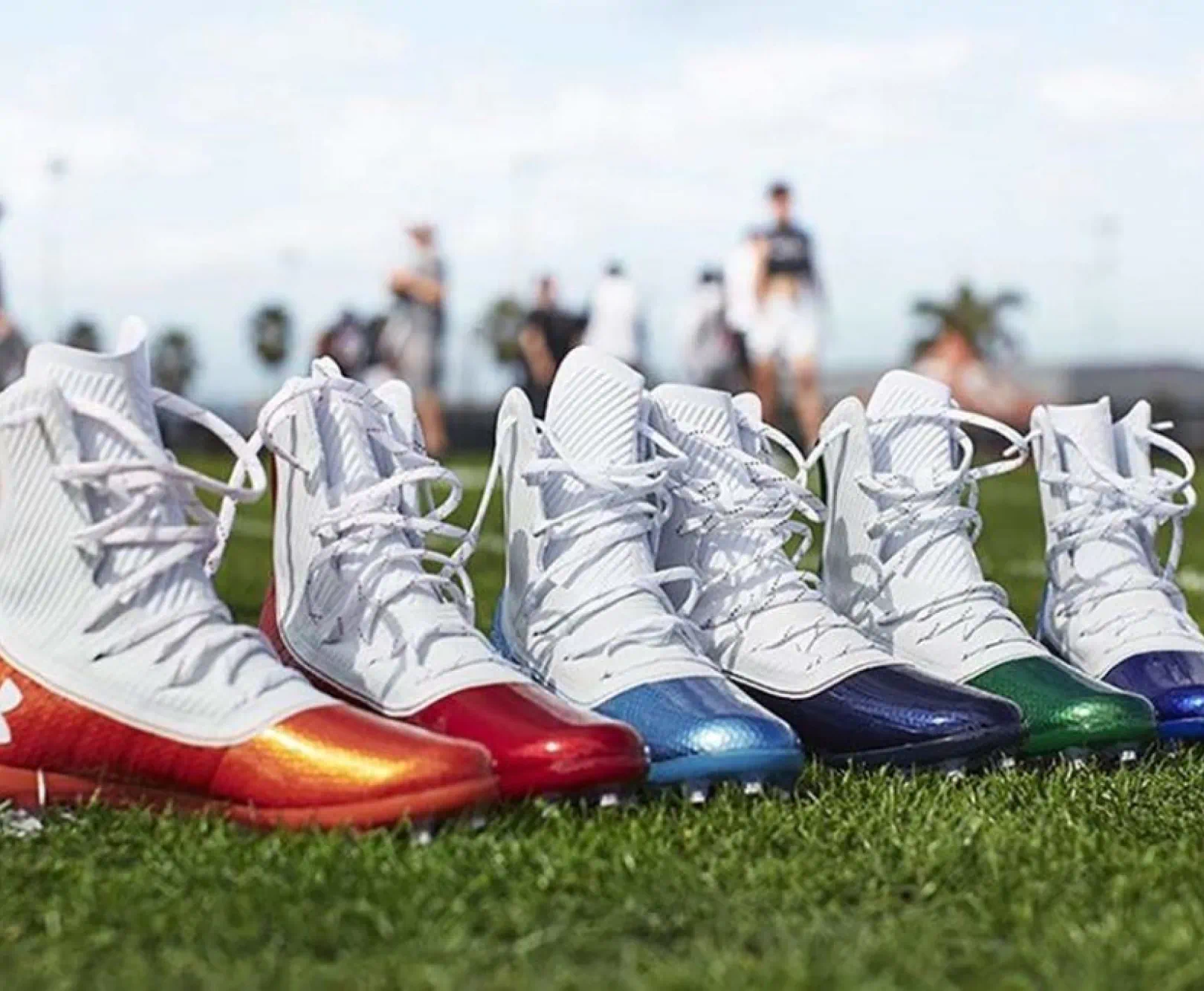 Several hightop football shoes sit upright on a bed of turf. Each with colorful toes ranging from orange to blue. Players practice in the background.